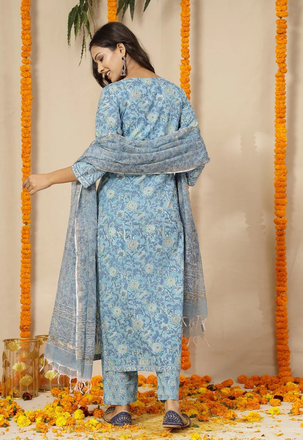 Woman in a blue floral traditional outfit standing against a decorated background with marigold flowers.