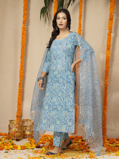 Woman in a light blue floral dress with a dupatta, standing against a decorated background with orange flowers and green leaves.